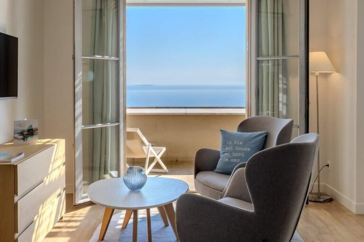 A modern room with a view of the sea, featuring a sofa, a small table with a vase, and a TV on a wooden cabinet.
