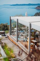 Un espace de restauration extérieur moderne avec vue sur la mer, doté de murs en verre, de planchers en bois et de mobilier d’extérieur.