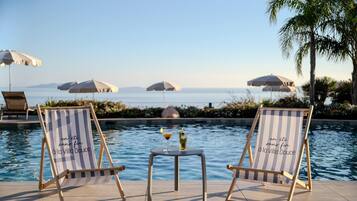 Twee gestreepte lounge fauteuils met een tafeltje en drankjes bij een zwembad, met parasols en uitzicht op zee.
