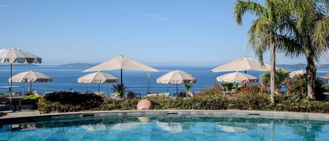 A pool with clear blue water, surrounded by umbrellas and palm trees, overlooking a body of water.