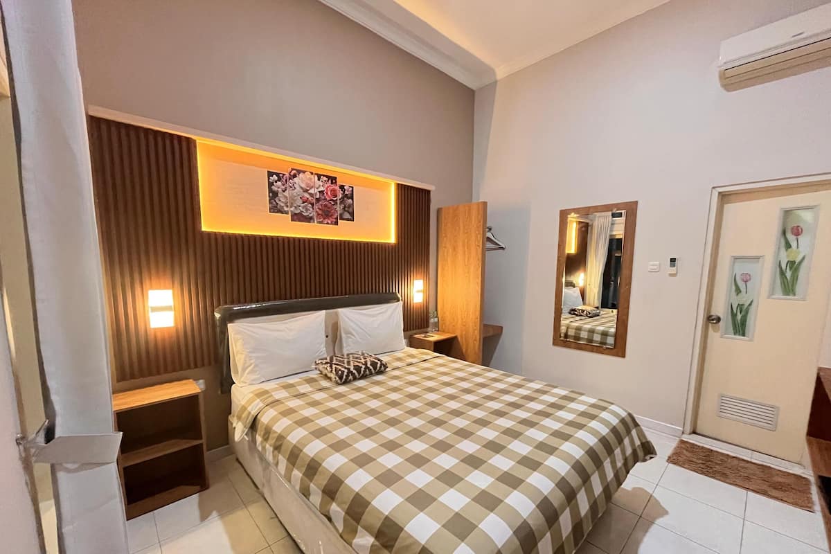 A hotel room with a checkered bedspread, a wooden headboard, a wall-mounted mirror, and a framed floral artwork above the bed.