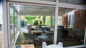 A glass-enclosed patio area with round tables and chairs, a small play area with a toy kitchen set, and a view of a brick building and greenery outside. - Hotel Alfa Inn (Blankenberge)