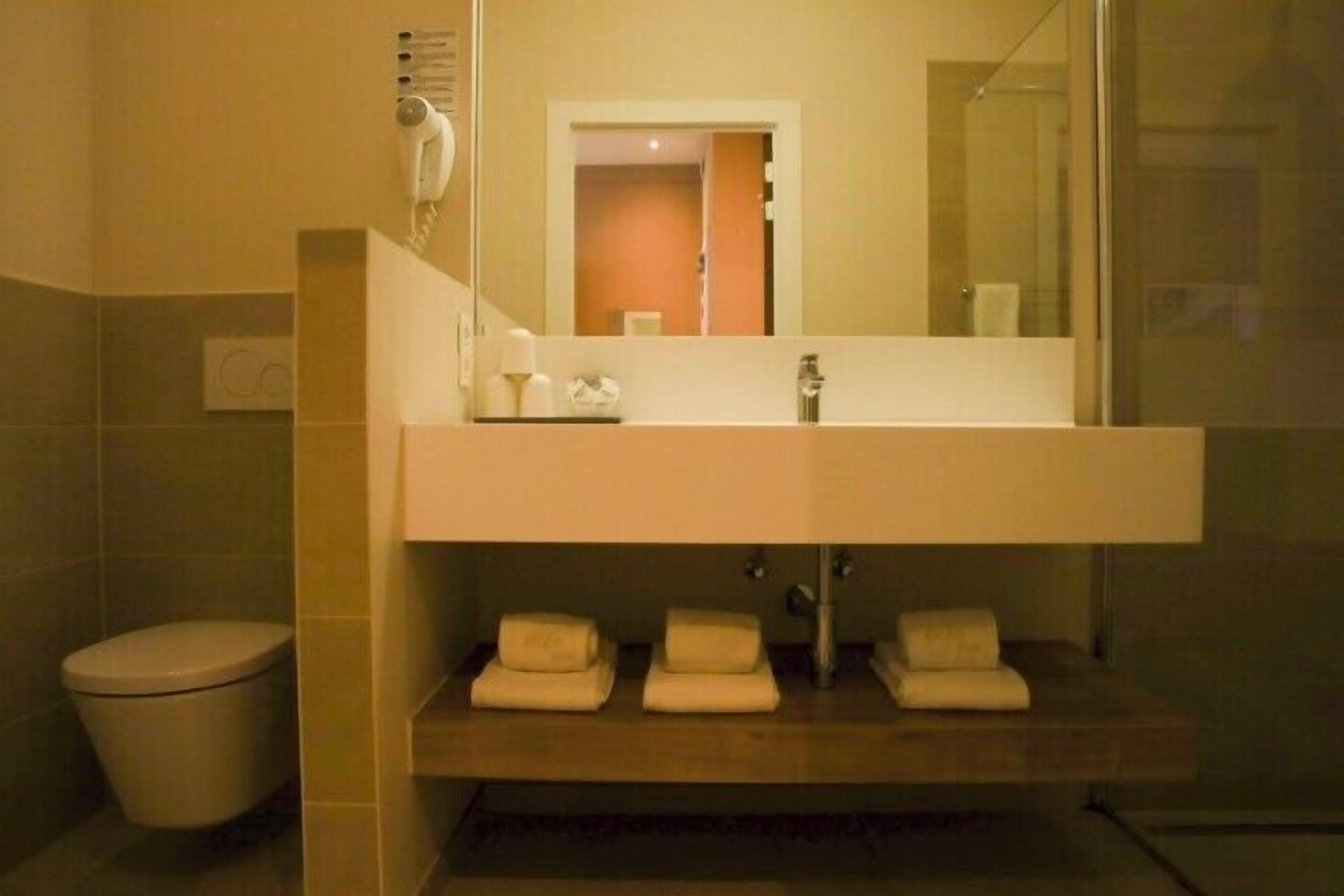 Une salle de bain avec un grand miroir, un lavabo et des toilettes. Des serviettes sont posées sur le plan du lavabo.