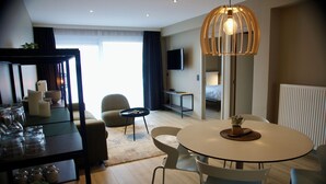 Una habitación de hotel moderna con una mesa de comedor redonda, un sofá, un televisor y un gran ventanal con cortinas.