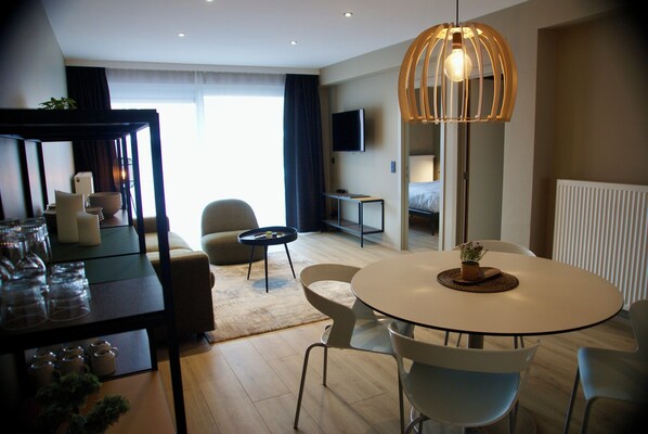 Une chambre d’hôtel moderne avec une table à manger ronde, un canapé, un téléviseur et une grande fenêtre avec des rideaux.