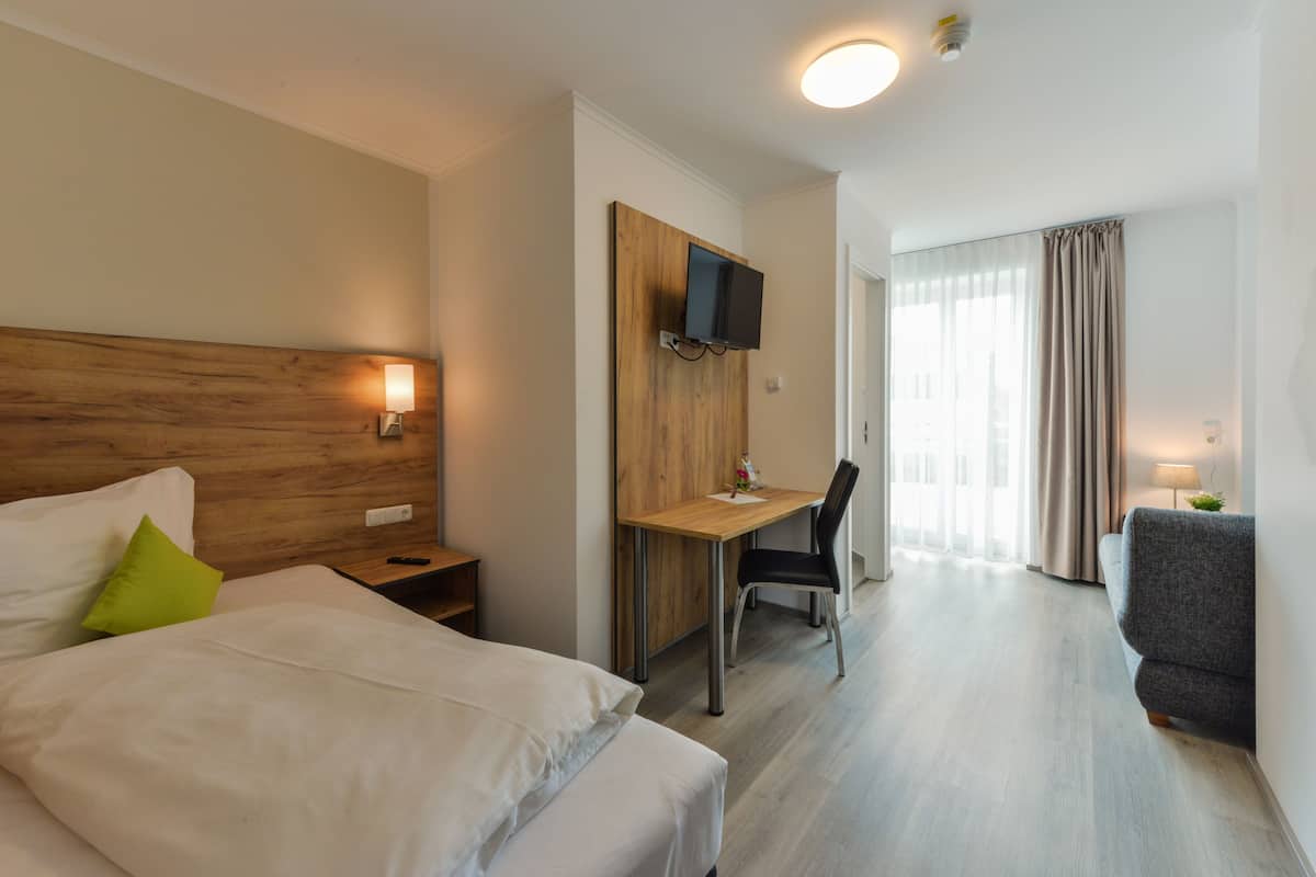 A hotel room with a bed, a desk, a chair, a TV mounted on the wall, and a window with curtains.