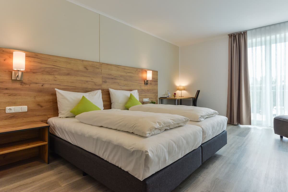 A modern hotel room with a large bed, bedside lamps, a nightstand, and a view of the city through large windows.