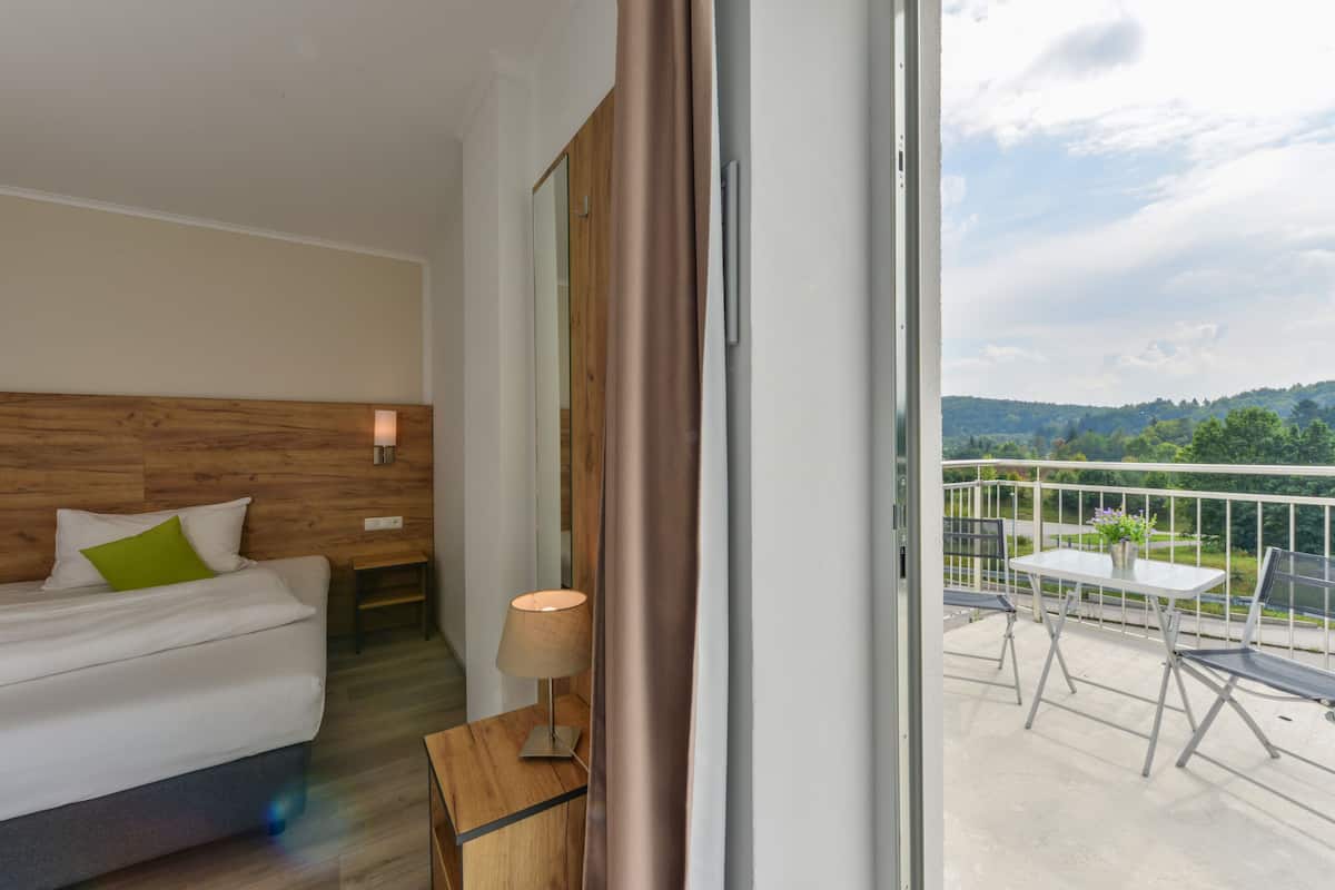 A modern hotel room with a wooden headboard, a bed with white bedding and a green pillow, a bedside table with a lamp, and a balcony with a table and chairs.