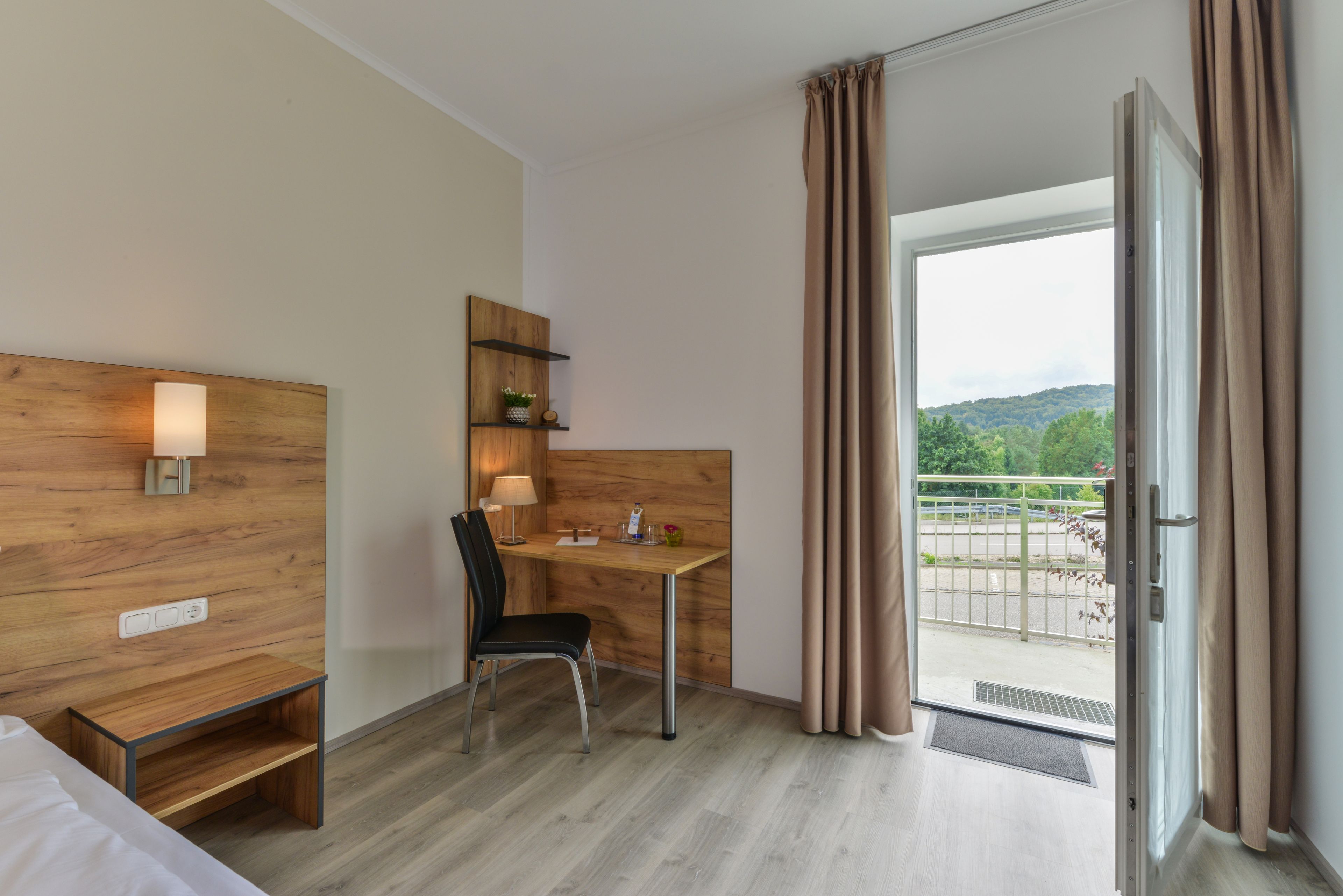 A hotel room with a wooden desk, chair, and bedside table. There is a window with curtains, a view of greenery outside, and a door leading to an outdoor area.