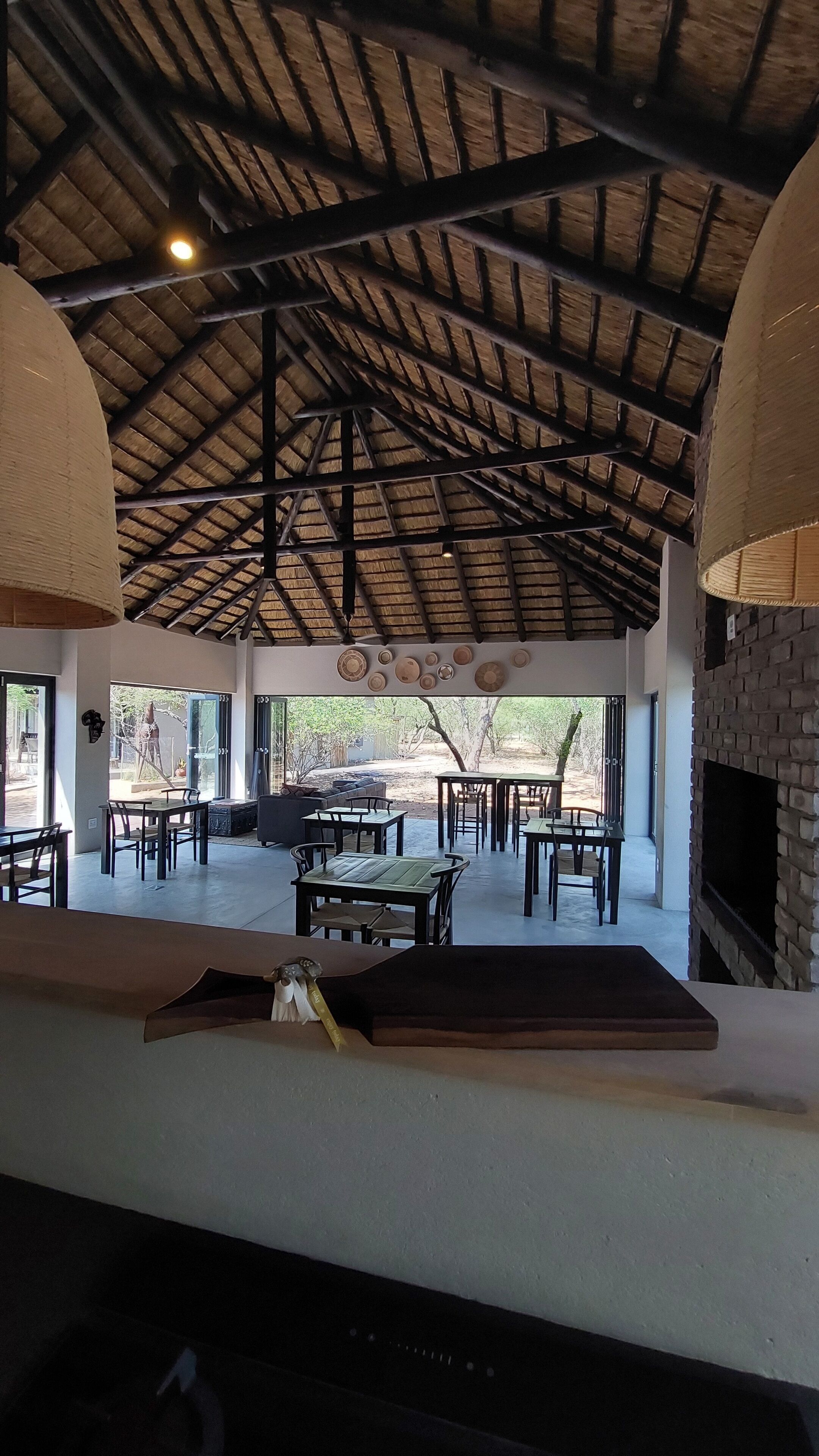 An indoor dining area with a high ceiling, wooden beams, and a brick fireplace. Tables and chairs are arranged for dining.