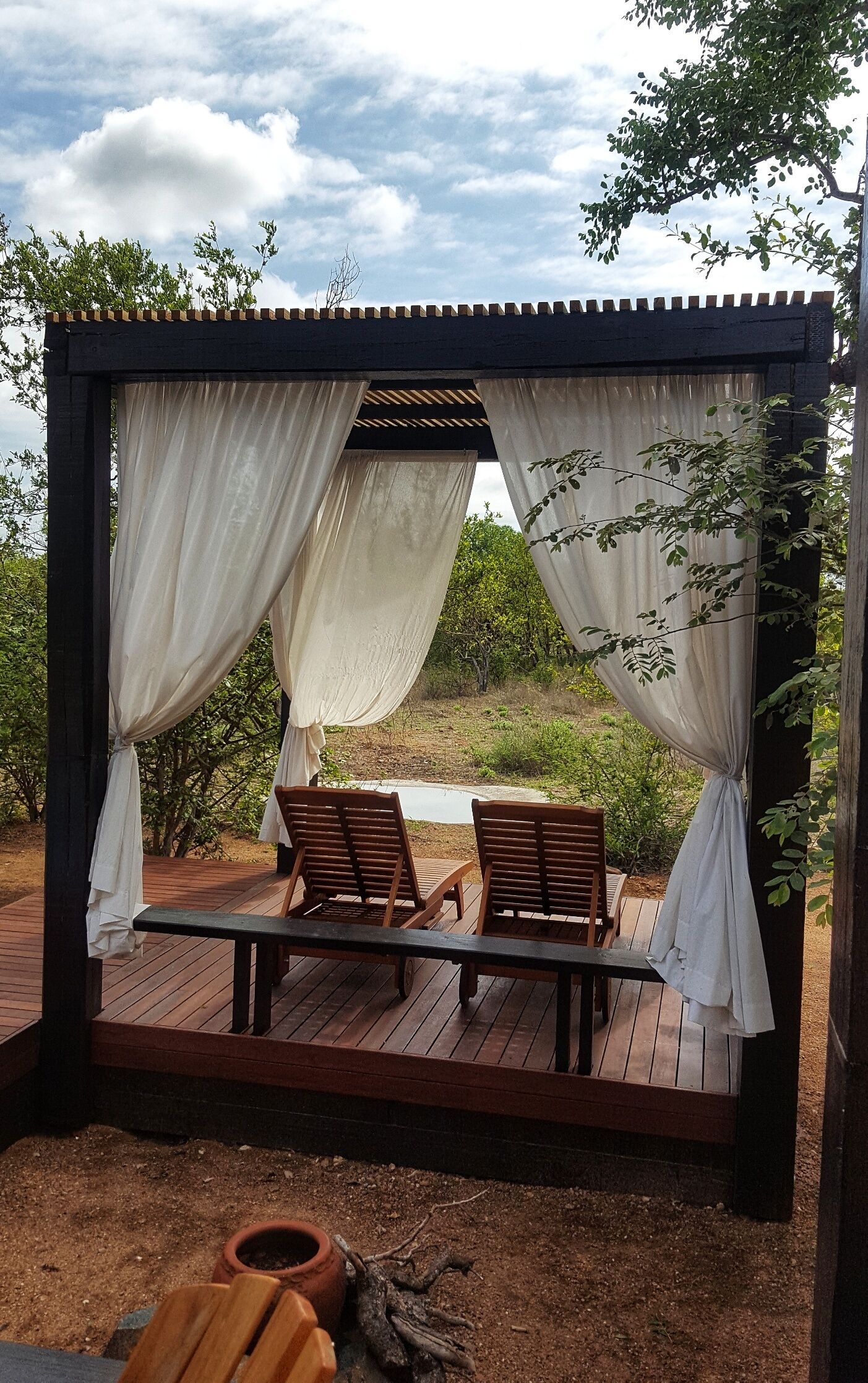 A wooden deck with two wooden chairs under a pergola with white curtains.