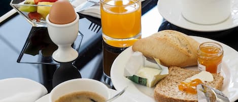 Mesa de café da manhã com diversos alimentos, incluindo torradas, geleia, manteiga, uma xícara de café, um copo de suco de laranja, uma tigela de frutas e um ovo pequeno.