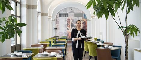 Une salle à manger d’hôtel avec des tables et des chaises, un serveur portant des plateaux et de grandes fenêtres.