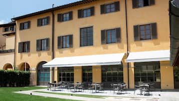 Un edificio con fachada amarilla, varias ventanas con contraventanas y zona de estar exterior con sombrillas.