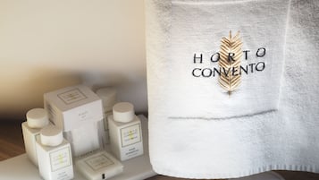 A hotel room with a neatly arranged bathroom vanity. On the vanity, there are several toiletries including a body lotion, a hair shampoo, and a body wash. Additionally, there are slippers with the hotel name embroidered on them.