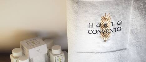 Un cuarto de hotel con una tocador de baño ordenado. En el tocador, hay varios artículos de tocador, incluyendo una loción corporal, champú y gel de baño. Además, hay pantuflas con el nombre del hotel bordado.