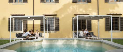 A pool area with two seating areas, each with a covered table and chairs, surrounded by a pool.