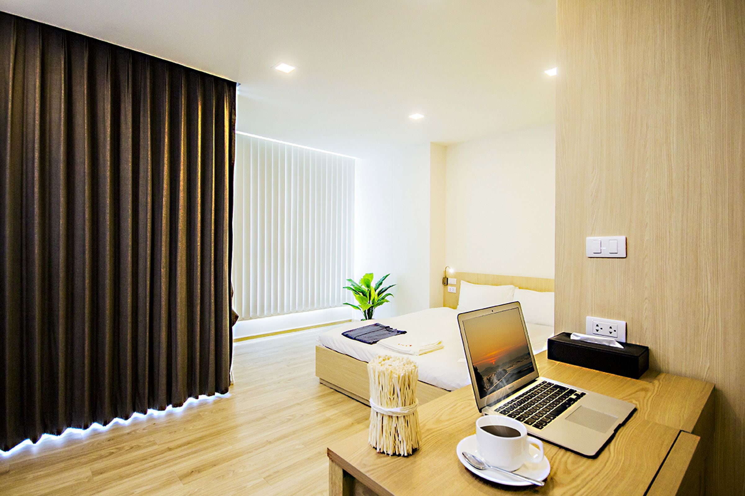 Una habitación de hotel moderna con un escritorio de madera, una computadora portátil, una taza y una mesita de noche con una planta.