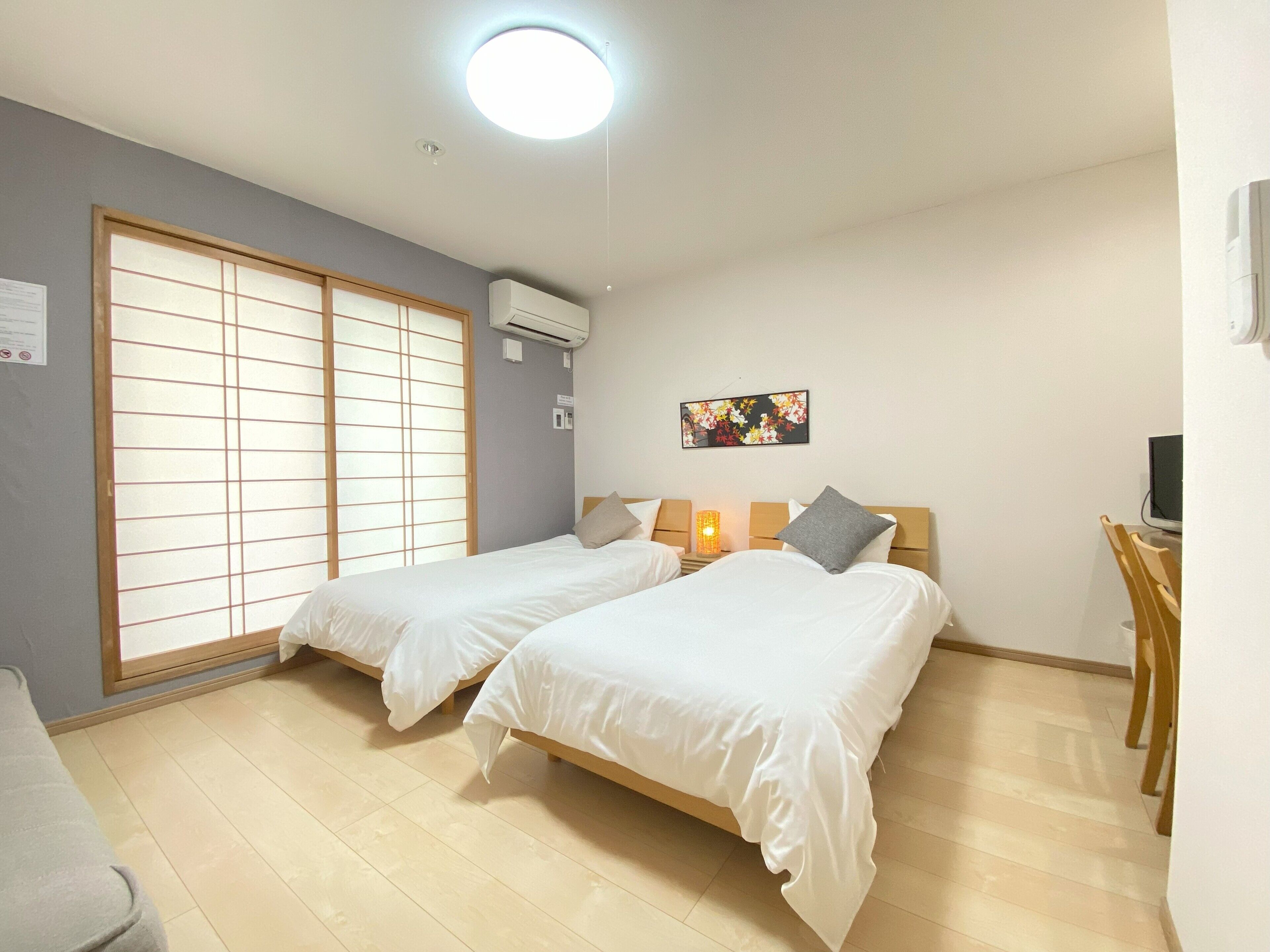 A hotel room with two beds, a large window with shoji screens, an air conditioner, a desk with a computer, and a small table with a lamp.