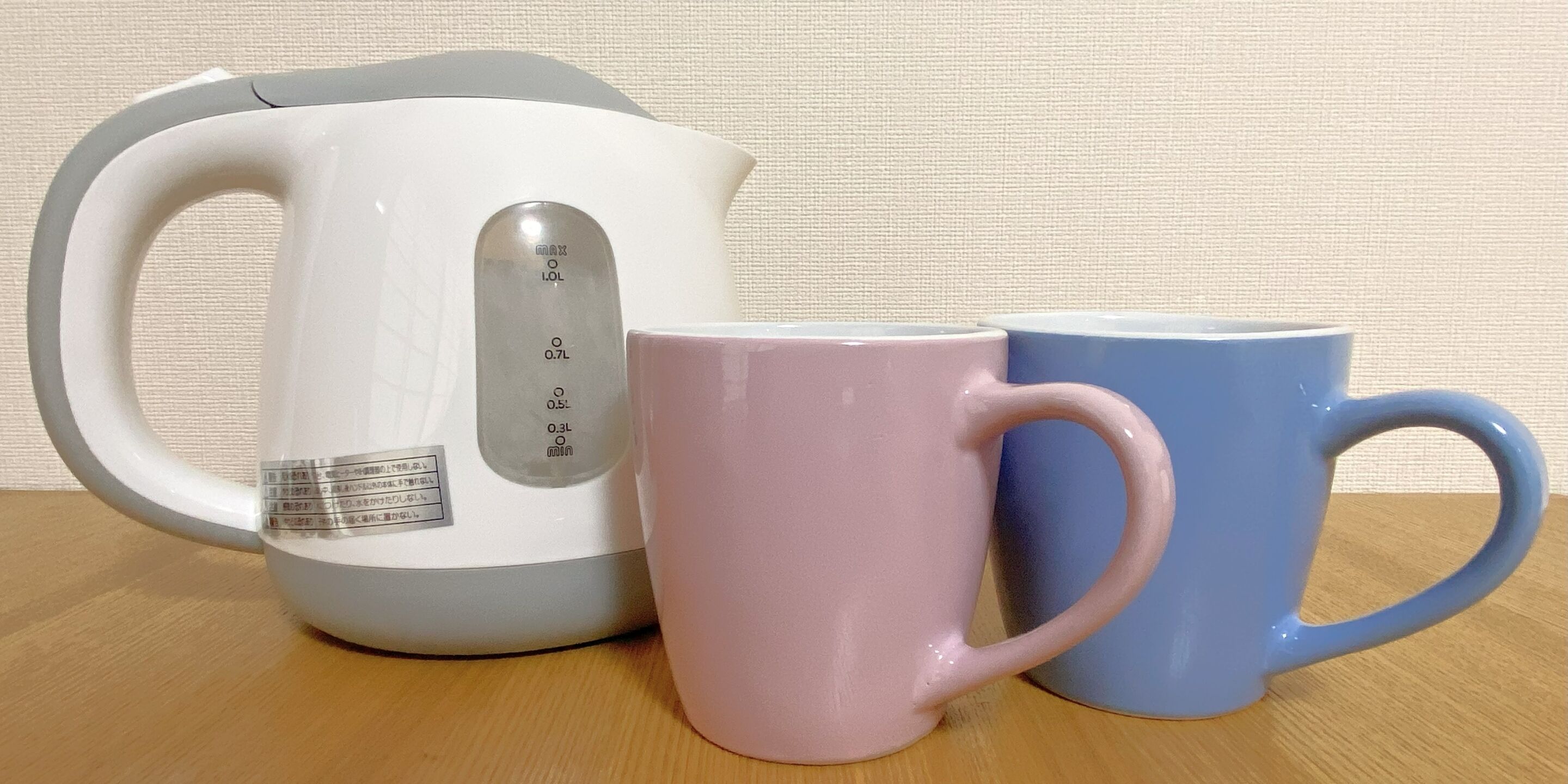A white kettle with measurement markings and a label, a pink mug, and a blue mug on a wooden surface.