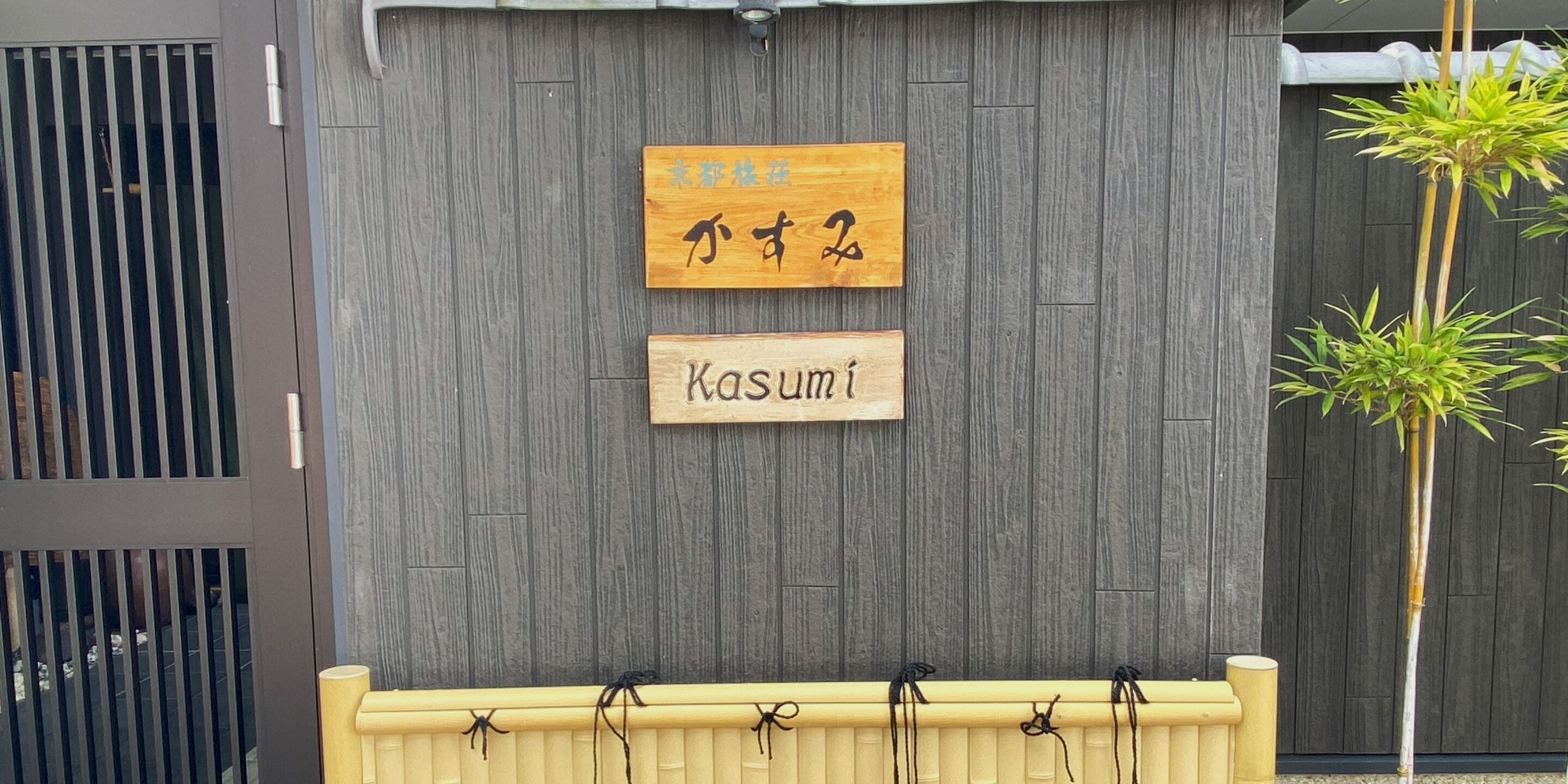 Un mur en bois sur lequel sont fixées deux enseignes : l’une porte des caractères chinois, l’autre le nom « Kasumi ».