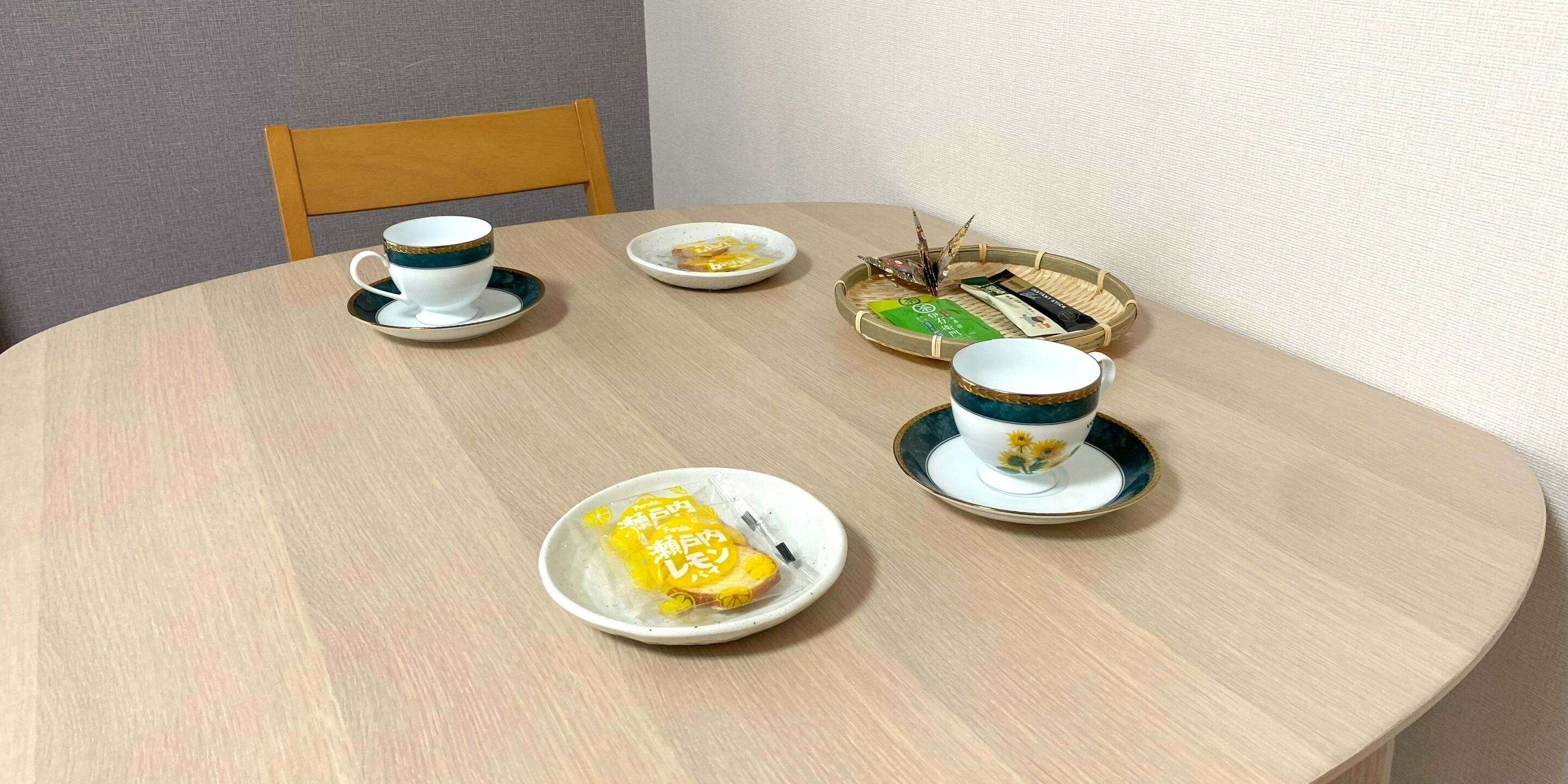 A round dining table with two white cups and saucers, a plate with a yellow item, and a basket with green and black items.