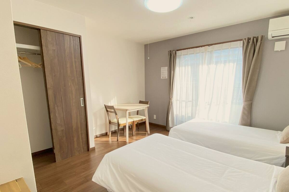 Habitación de hotel con dos camas, un escritorio y una silla. Cuenta con una ventana con cortinas, un equipo de aire acondicionado y una lámpara en la mesita de noche.
