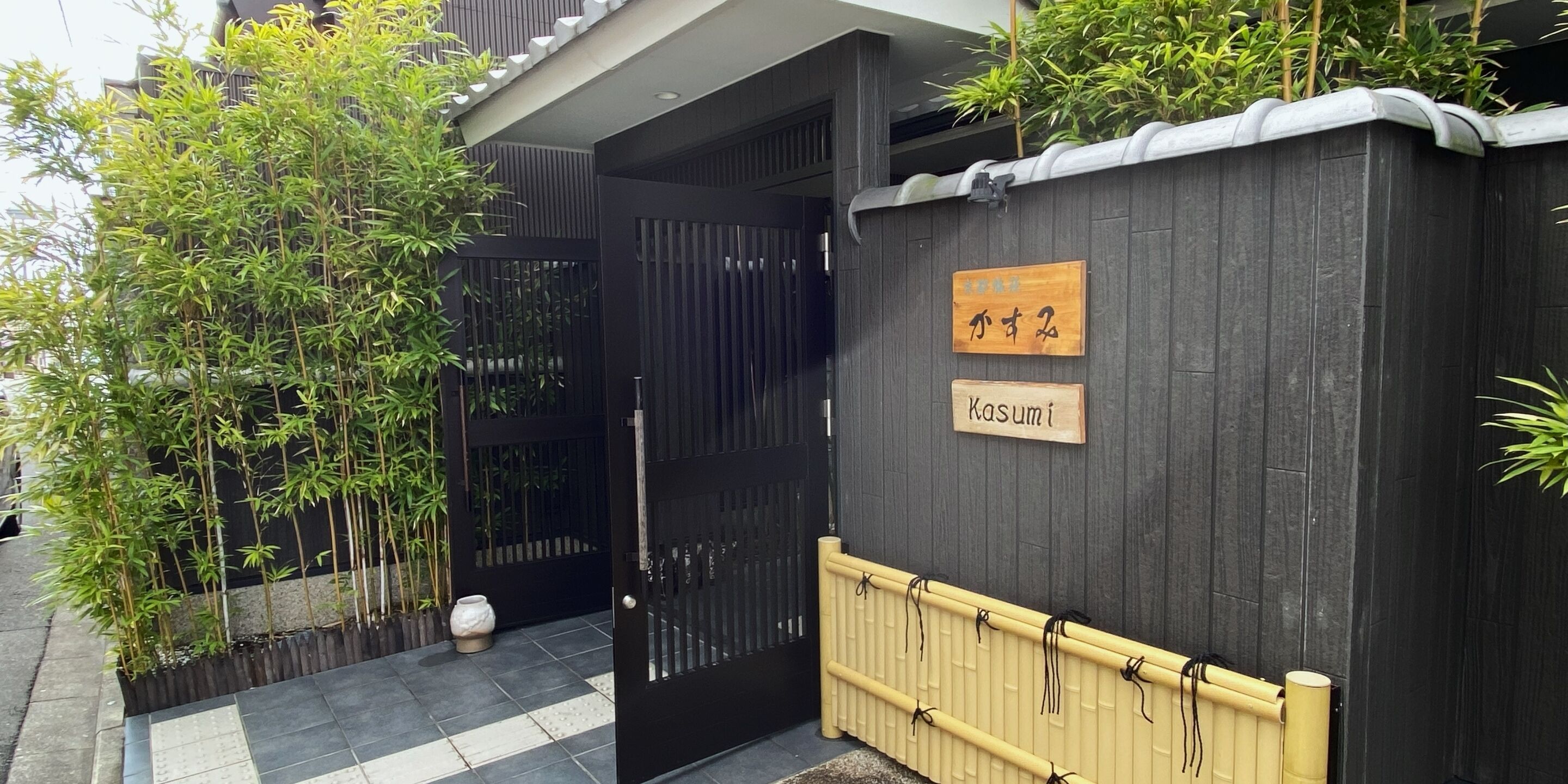 Een modern gebouw met een zwart hekwerk, een houten bord met Japanse karakters, en een bamboeplant ernaast.
