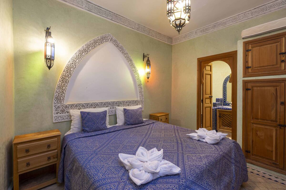 A bedroom with a large bed, a wooden nightstand, a wardrobe, and a bathroom visible through an arched doorway.