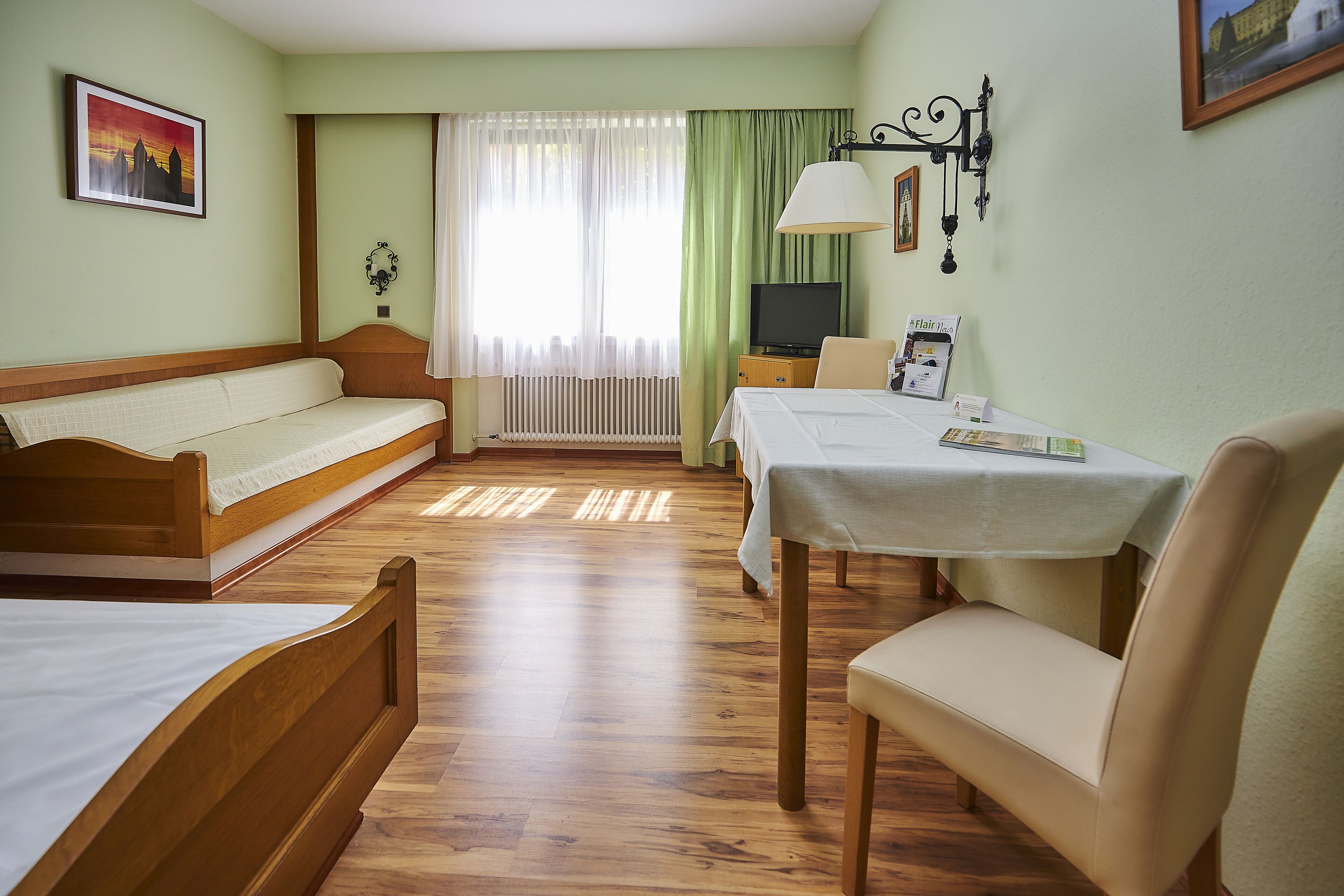 A hotel room with a wooden bed, a sofa, a dining table with a chair, a television, and a window with curtains.
