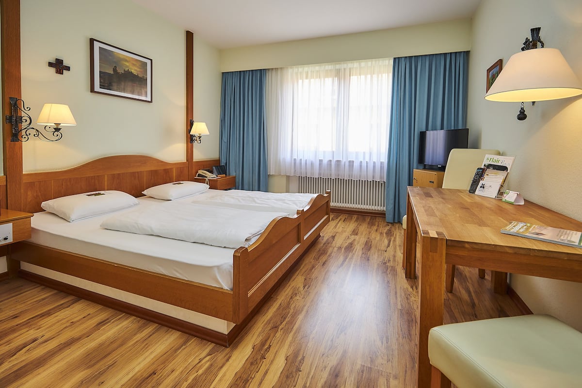 Une chambre d’hôtel avec un lit en bois, un bureau, une chaise, une télévision et une fenêtre avec des rideaux.