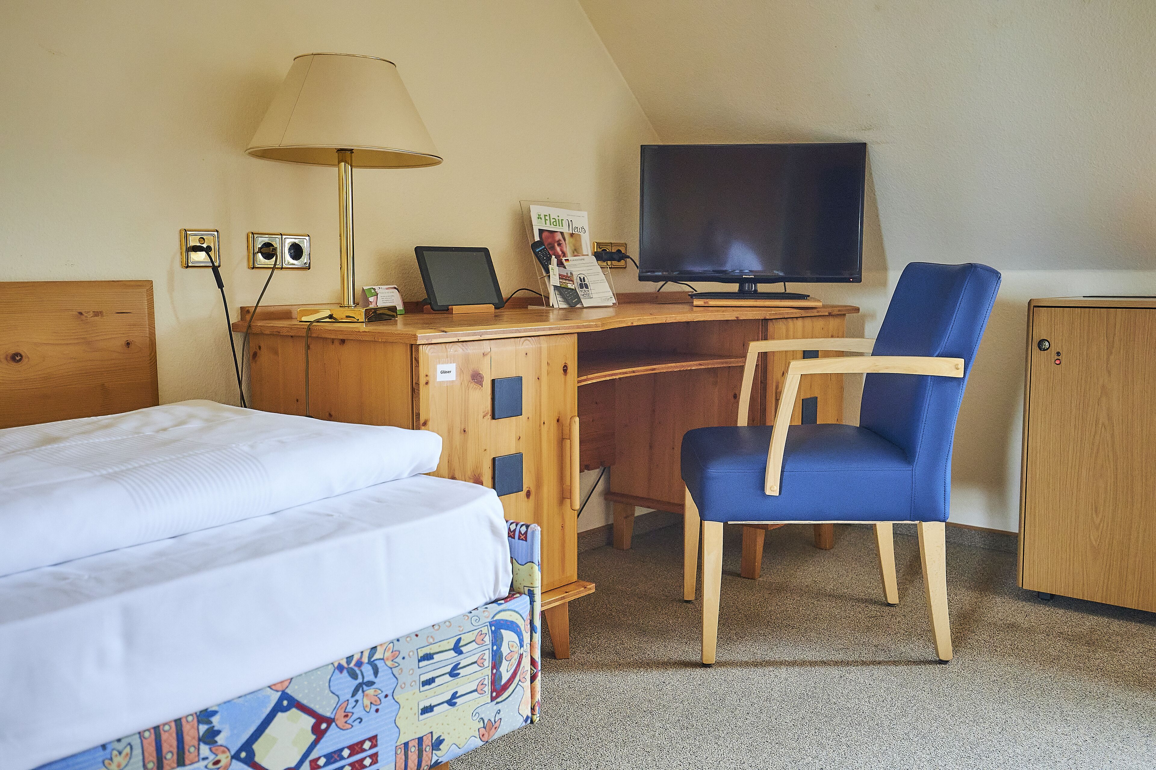 Quarto de hotel com cama, escrivaninha com computador, cadeira, televisão e armário.