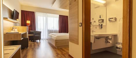 A hotel room with a wooden floor, a bed, a desk with a chair, a TV, a desk lamp, a bathroom with a sink, mirror, and toilet paper.
