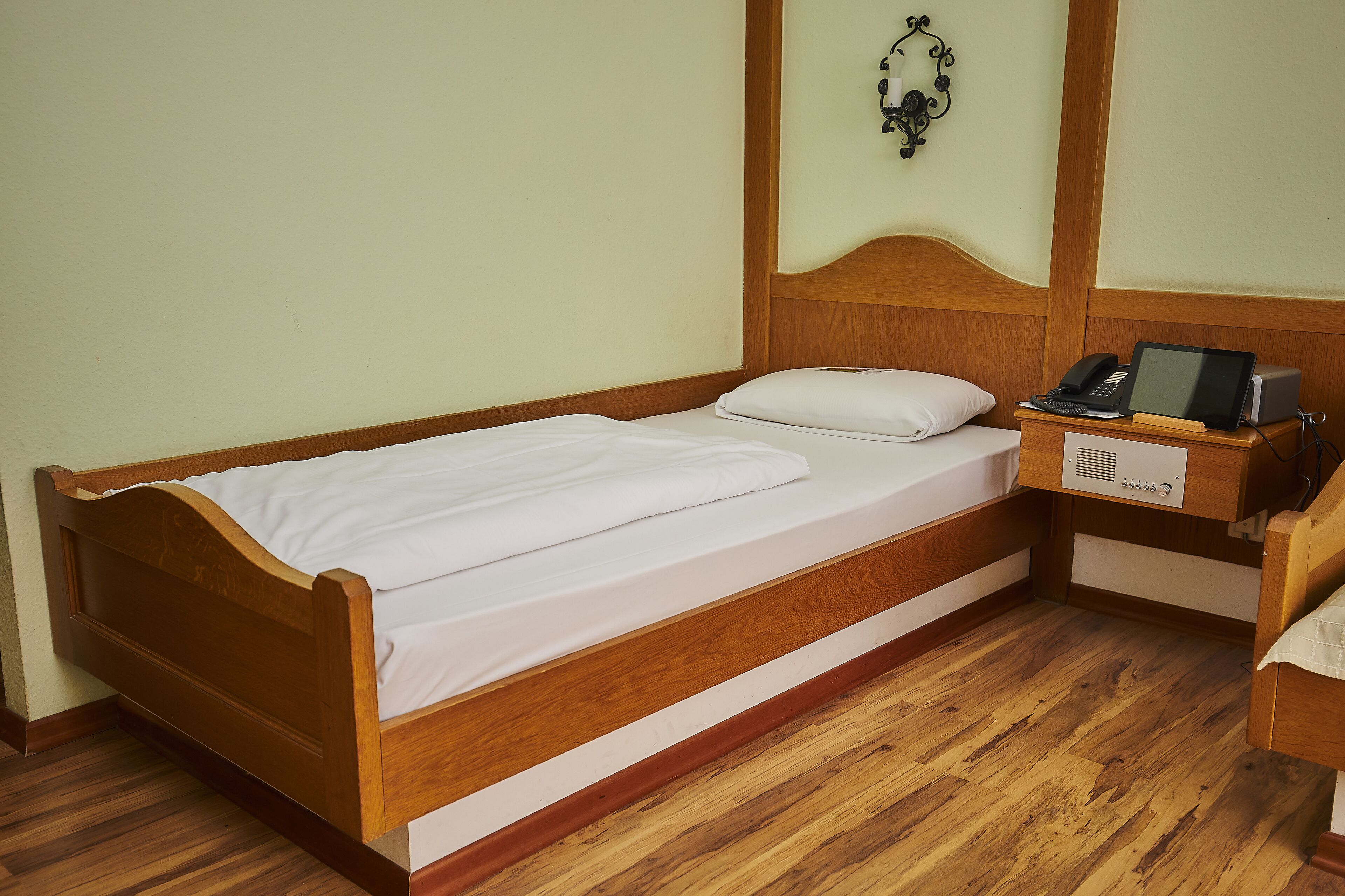 A single wooden bed with white bedding, a bedside table with a telephone and a book, and a wall-mounted light fixture.