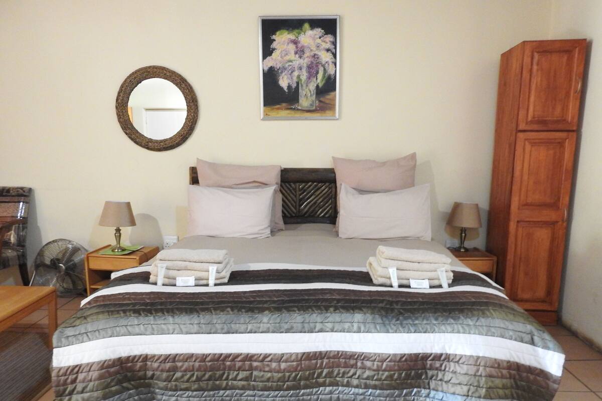 Uma cama bem arrumada com colcha listrada, duas mesas de cabeceira com luminárias, um guarda-roupa de madeira, um espelho redondo e um quadro com flores na parede.