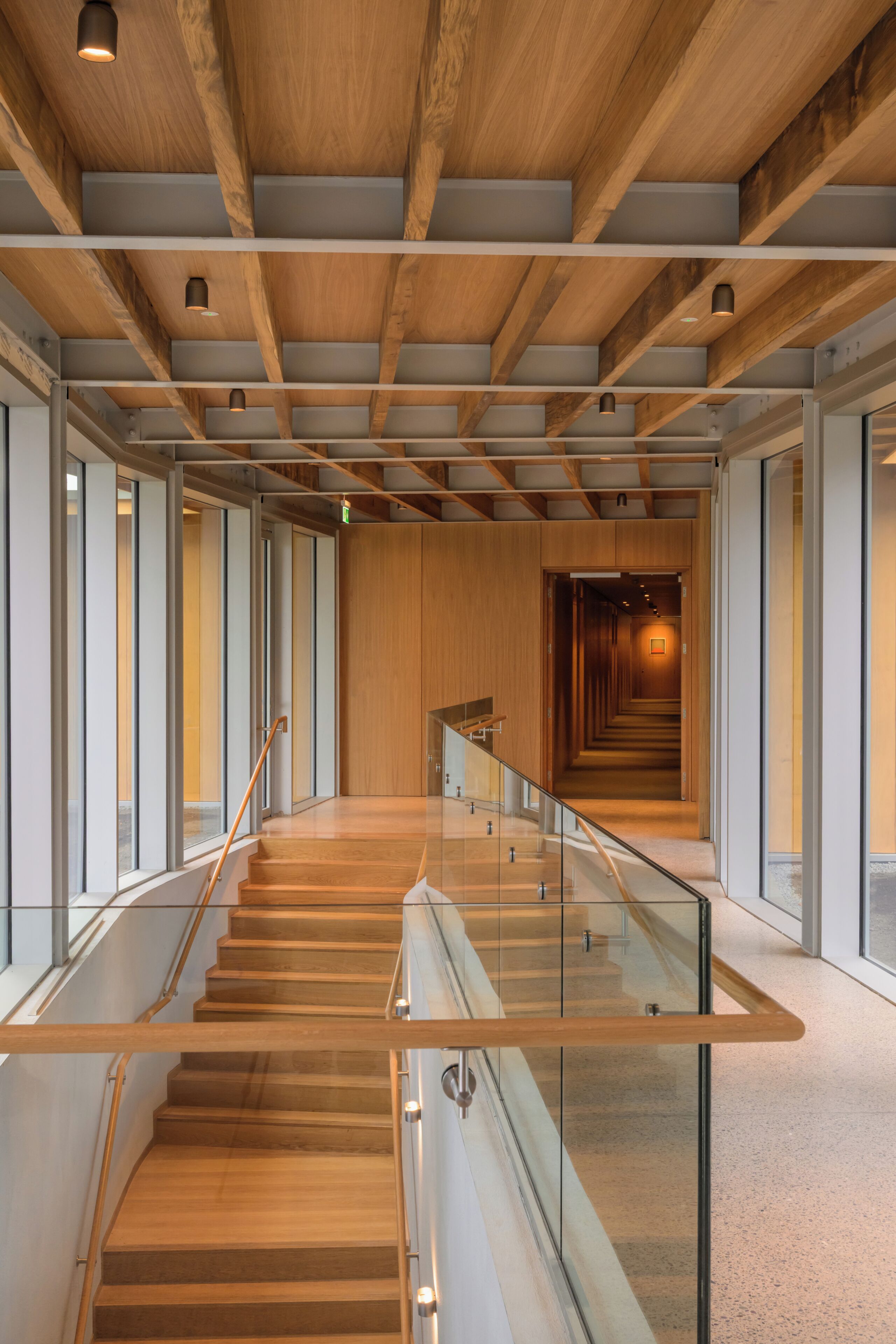 Un interior moderno con una escalera de madera, barandillas de cristal y un largo pasillo que conduce a una habitación con un cuadro.