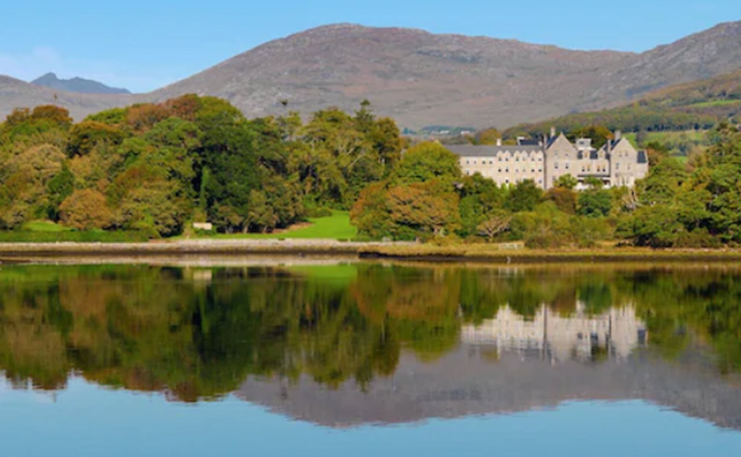 A large, historic building with multiple towers, situated by a calm lake with a mountain backdrop.
