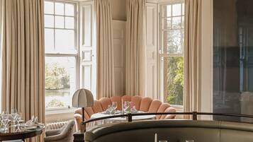 A dining area with a round table set for two, a cushioned bench, and a view of the outdoors through large windows.
