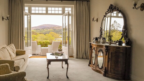 Habitación con sofá, sillas, una mesa y un espejo grande. Se ve follaje y colinas a través de las ventanas.