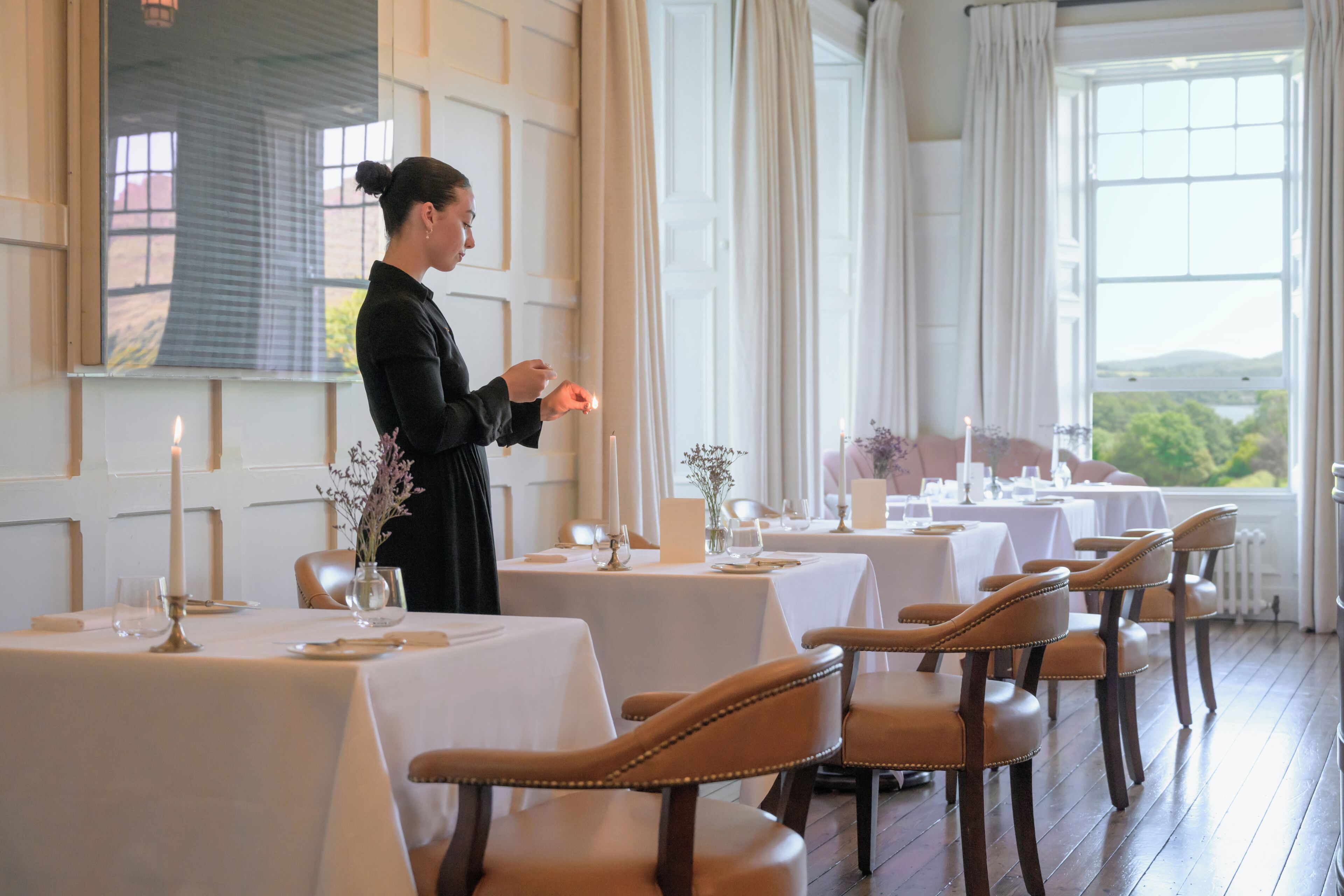 A dining area with tables set for a meal, a person attending to the tables, and a view of greenery outside.