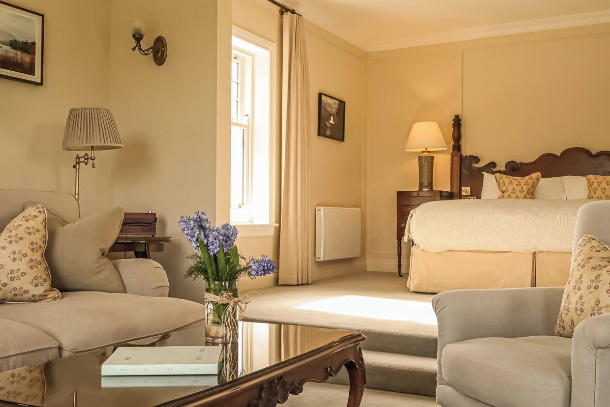 A hotel room with a bed, two armchairs, a sofa, a small table with a vase of flowers, a lamp, and a picture on the wall.