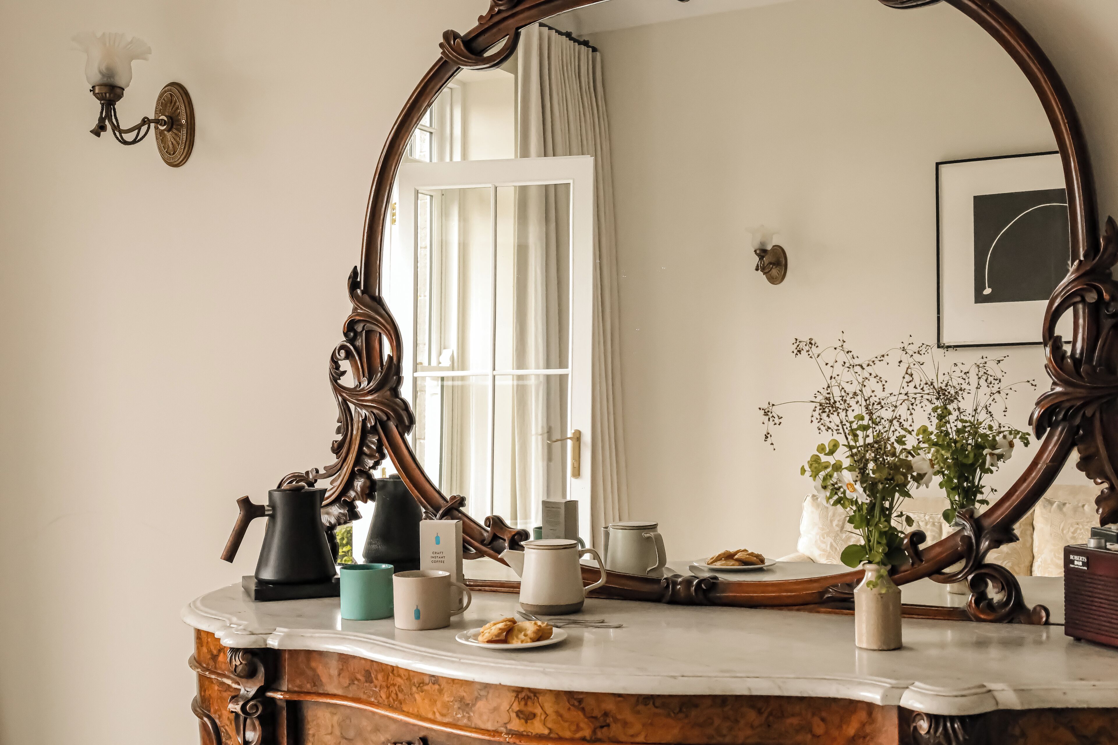 A vintage-style bathroom with a large ornate mirror, a marble countertop, and a window with curtains.
