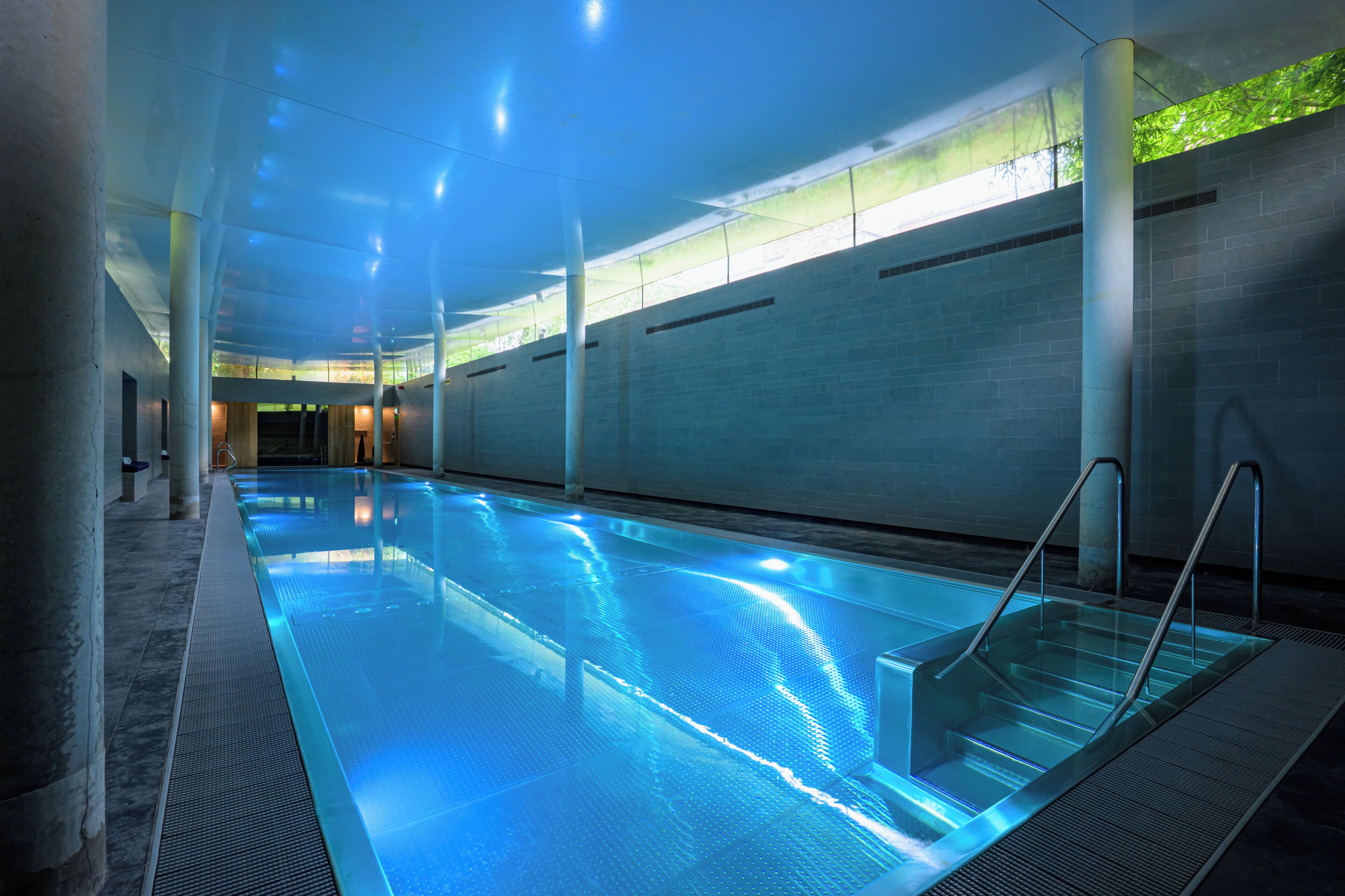 An indoor swimming pool with clear water, surrounded by walls and columns, and a glass ceiling allowing natural light.