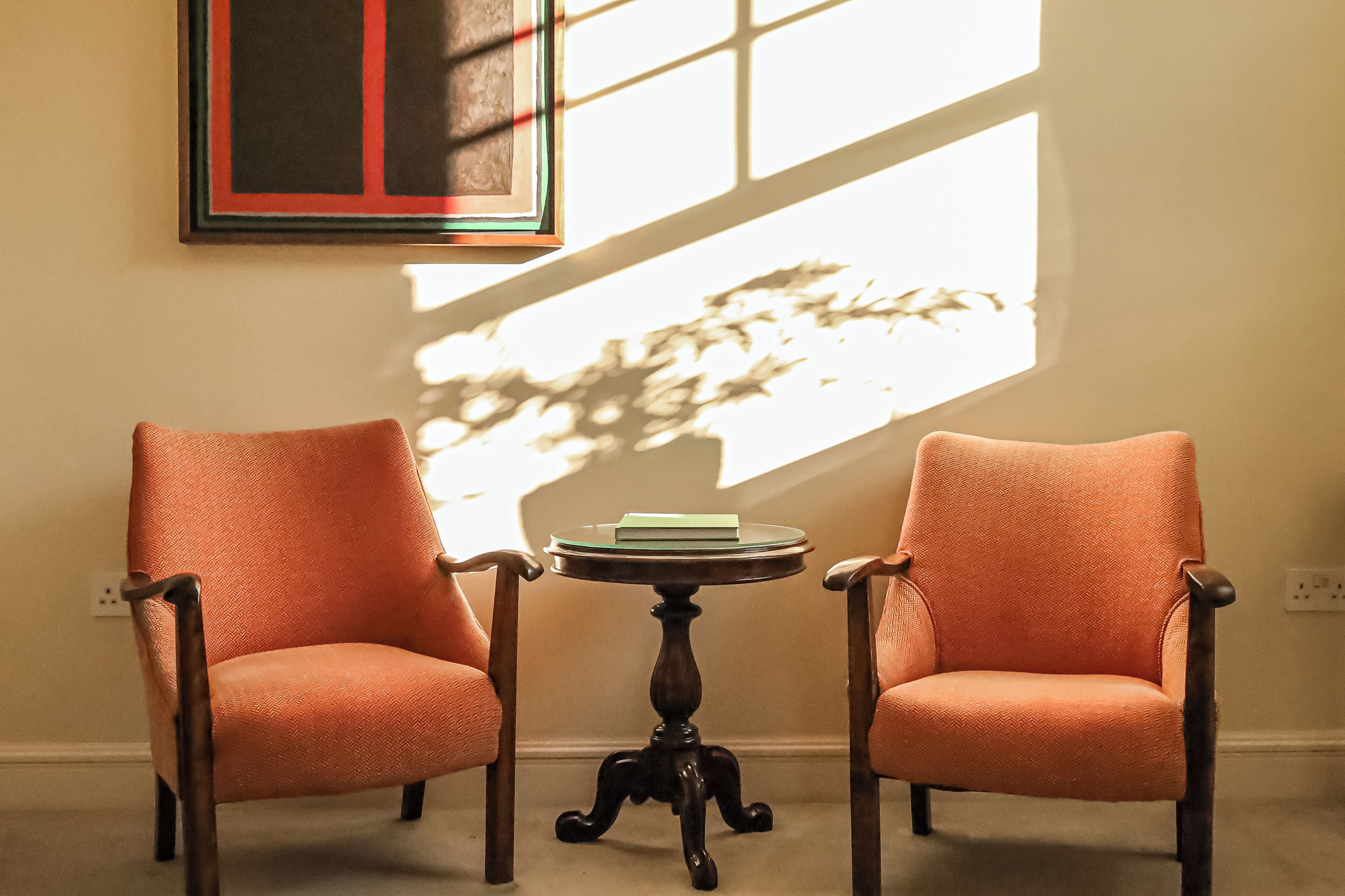 Two orange chairs with a small round table between them, a framed picture on the wall, and sunlight streaming through a window.