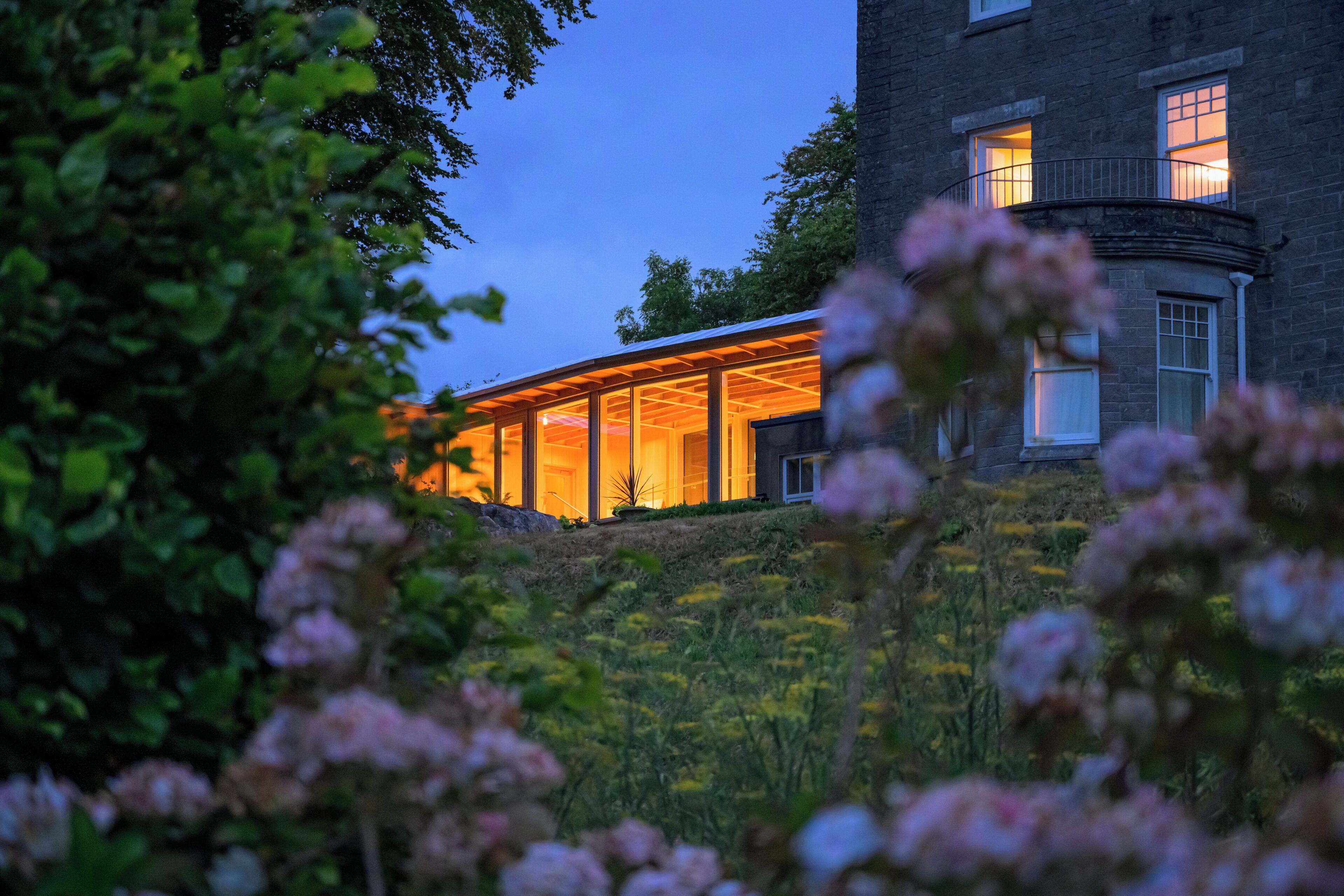A modern house with a glass facade, surrounded by greenery and flowers.