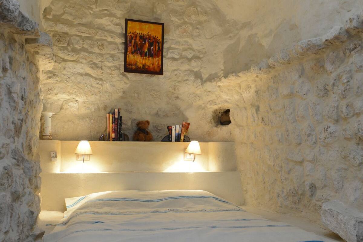 Un dormitorio con pared de piedra, una cama con colcha a rayas blancas y azules, un tapete rojo estampado y dos lámparas fijadas a la pared.