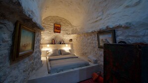 A cozy room with a stone wall, a bed with pillows, a nightstand with a lamp, and framed pictures on the wall. - L'Auberge De Tamezret (Matmata)
