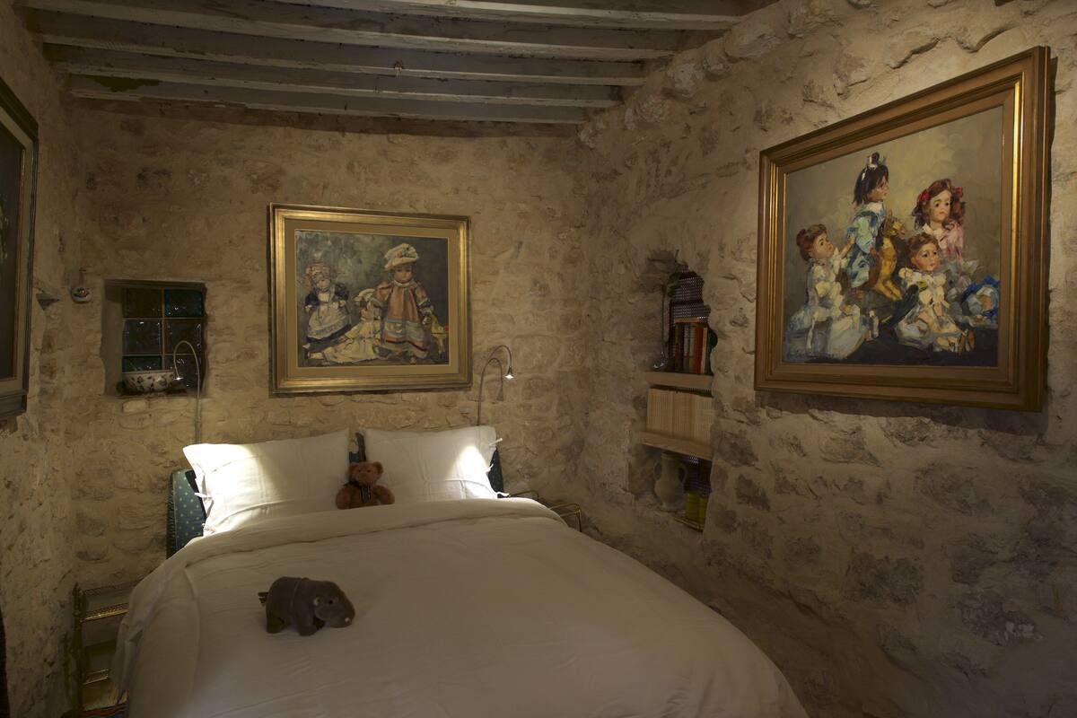 Un dormitorio con pared de piedra, una cama con ropa blanca, un osito de peluche y dos cuadros enmarcados.