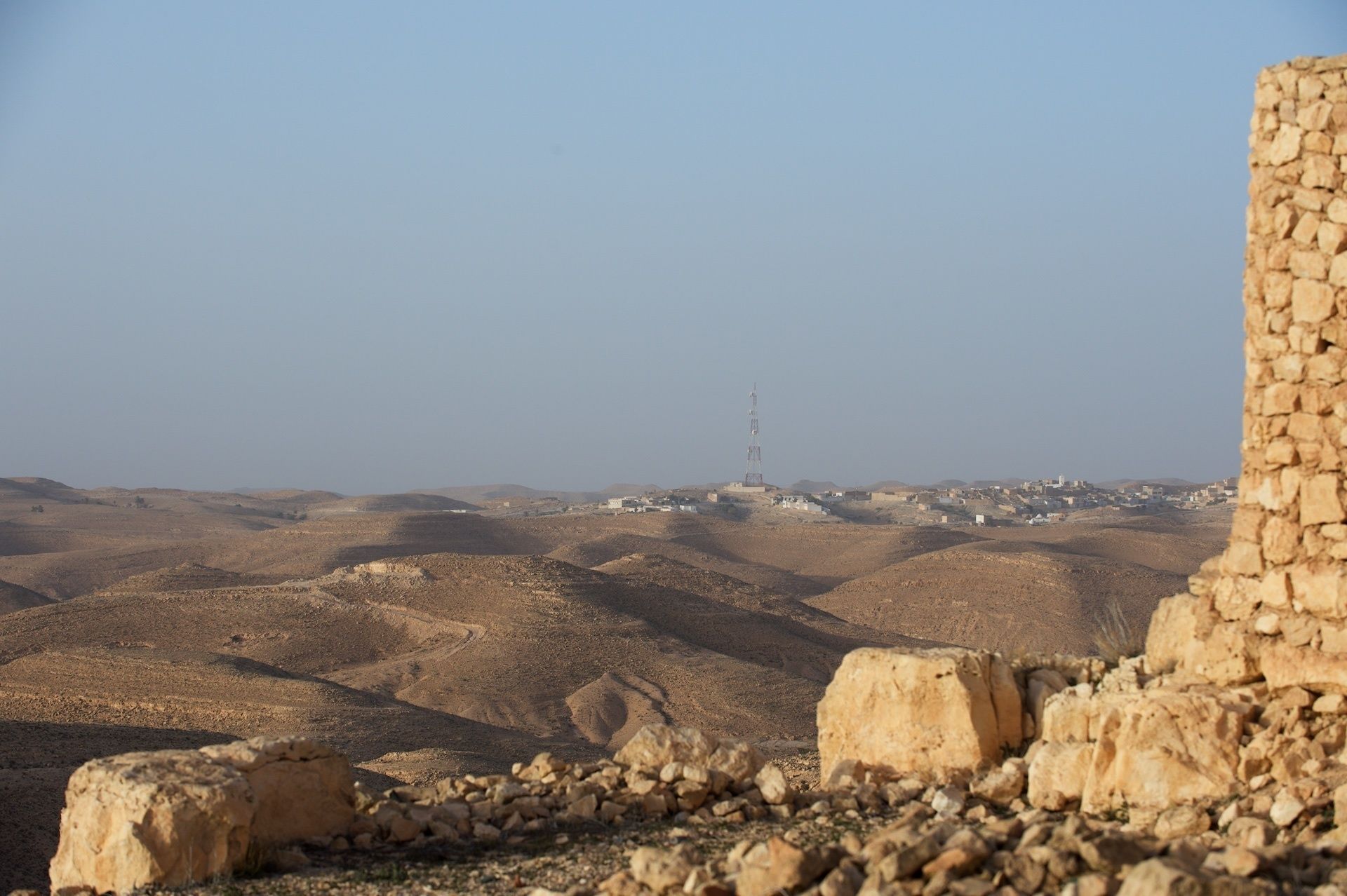 A desert landscape with a prominent communication tower in the distance and ancient stone ruins in the foreground.