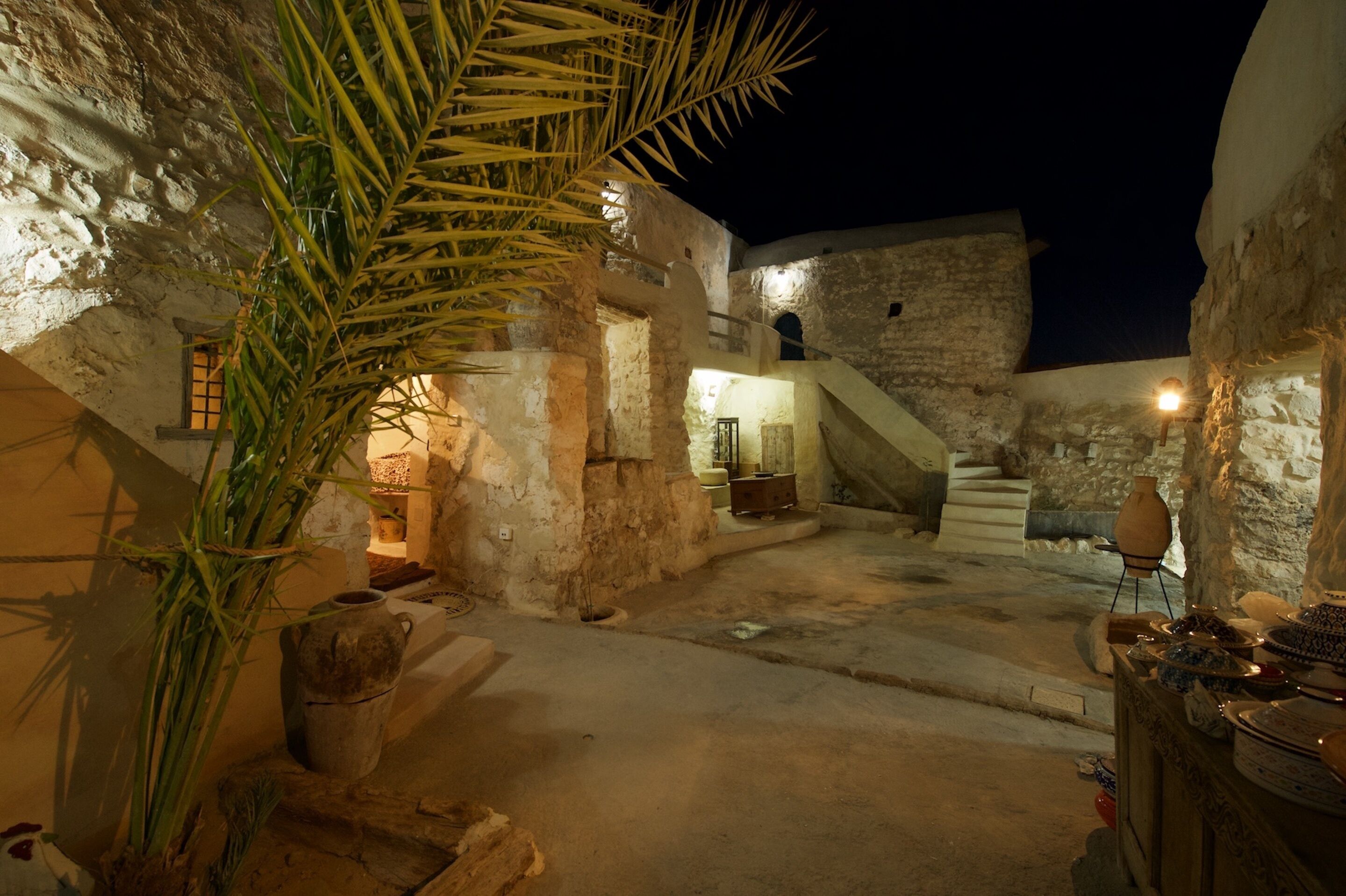 Un patio tenuemente iluminado con muros de piedra, una palmera y una escalera que conduce a un nivel superior.